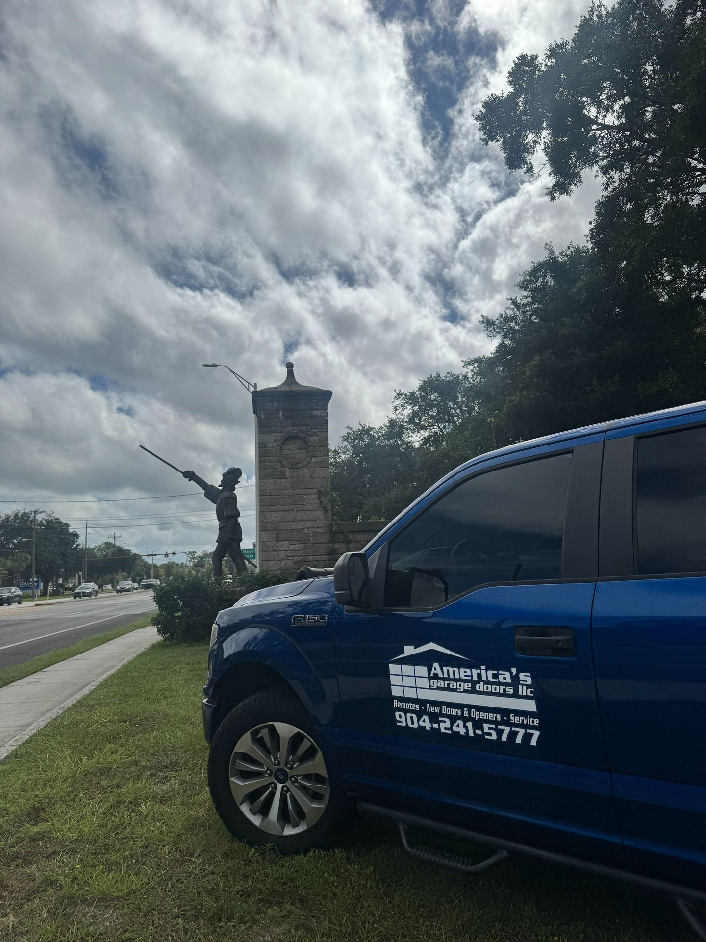 A bright blue truck parked on a grassy roadside near a tall stone pillar topped with a statue of a figure pointing up.