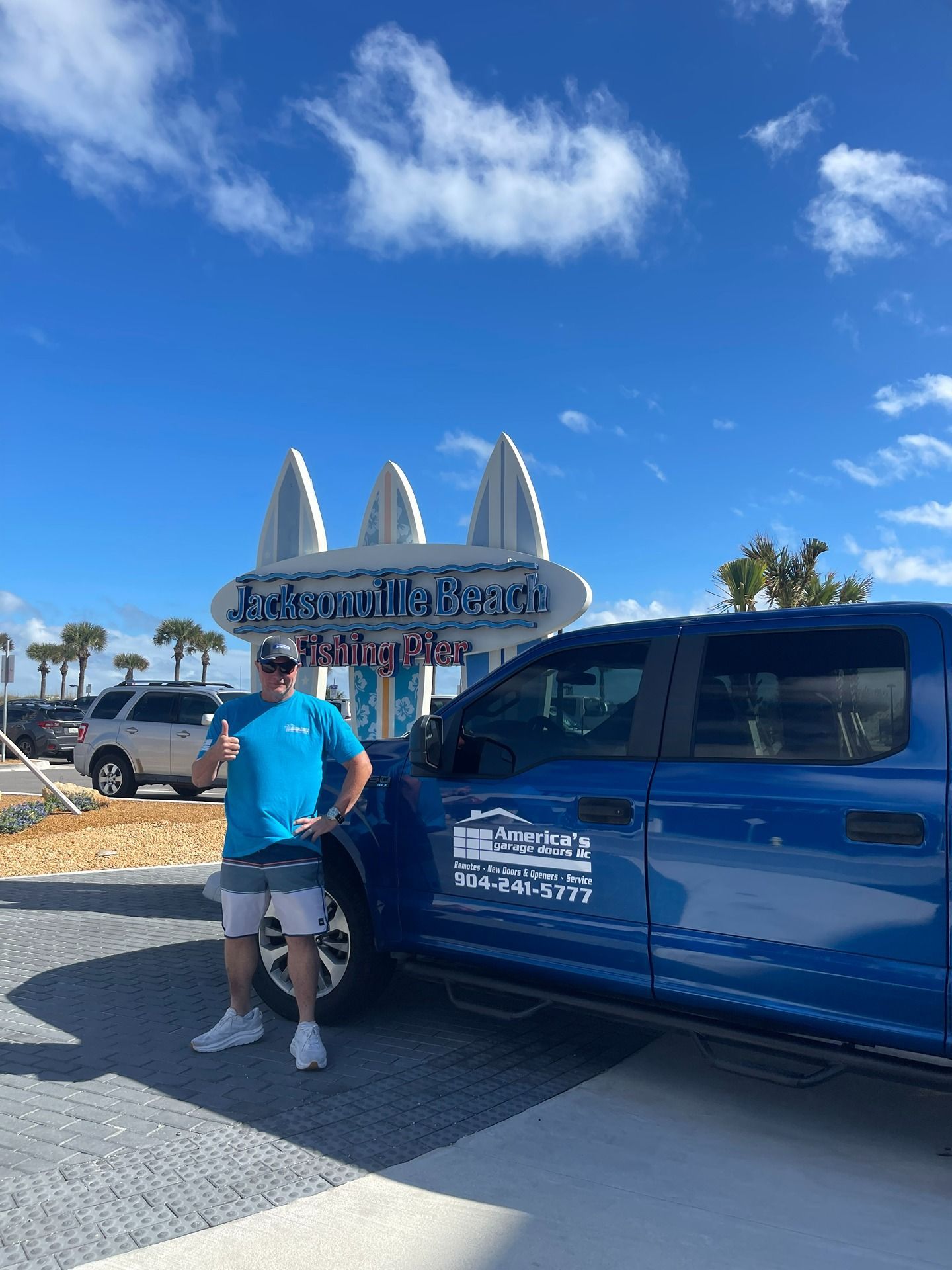 A person wearing a blue shirt stands next to a blue truck in front of a Jacksonville Beach surfing sign.