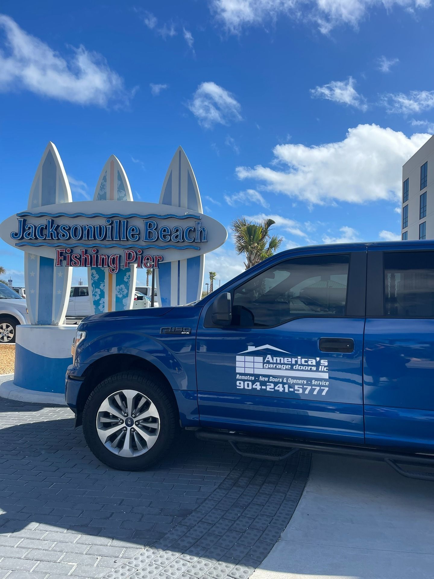 A blue pickup truck parked in front of the Jacksonville Beach Fishing Pier sign under a blue sky with white clouds.