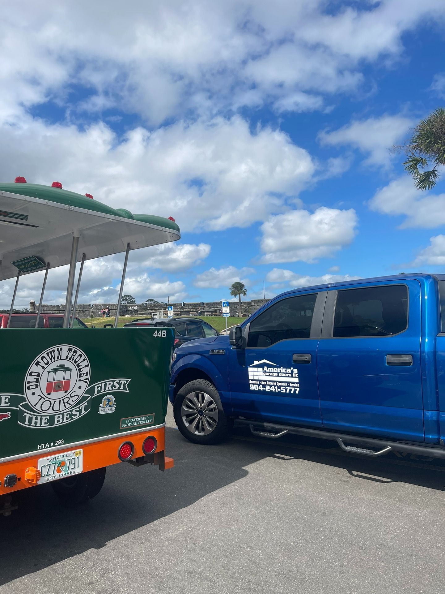 A blue pickup truck parked next to a green and orange sightseeing trolley against a blue sky with clouds.