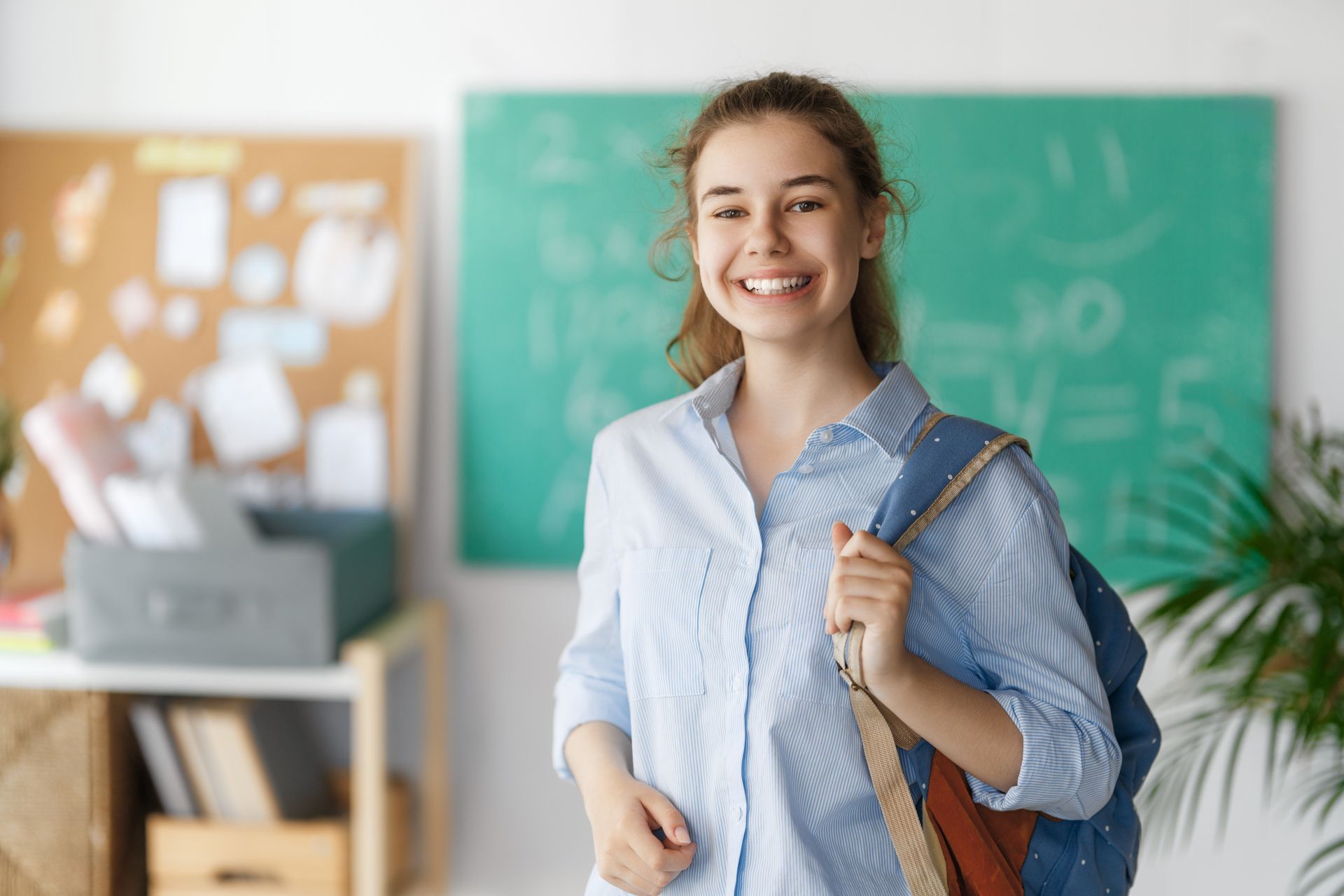 A young woman with a backpack is smiling in front of a green board.