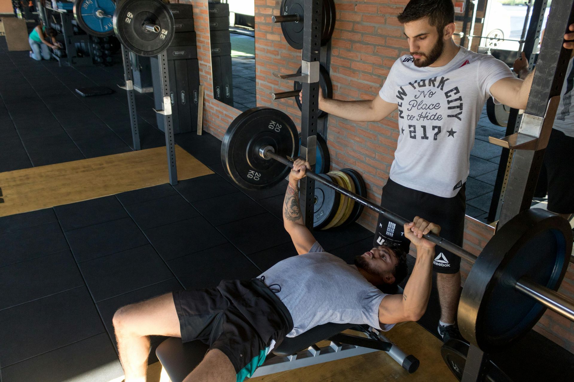 A man standing over another man who is laying on a bench doing a chest press with a barbell