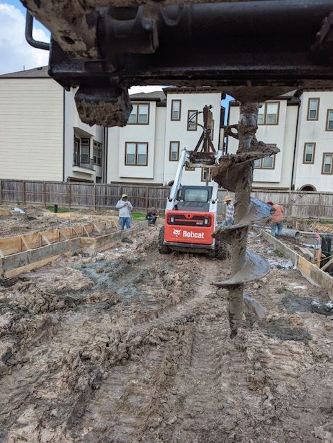 Construction site with auger drilling, Bobcat, muddy ground, workers, houses in the background.