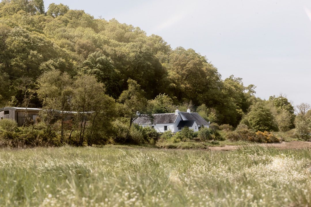 A white cottage with a dark roof nestled beneath a green hillside, tall grass in foreground.