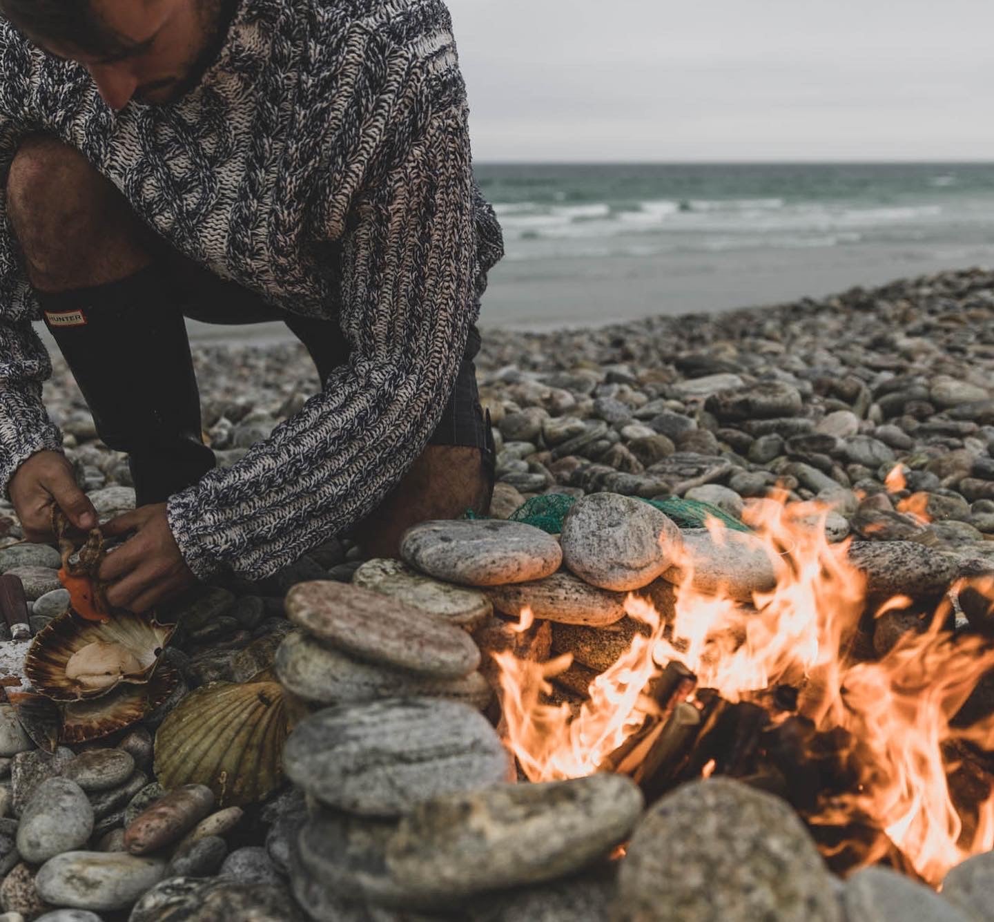 Chef preparing scallops over an open fire on a pebble beach.