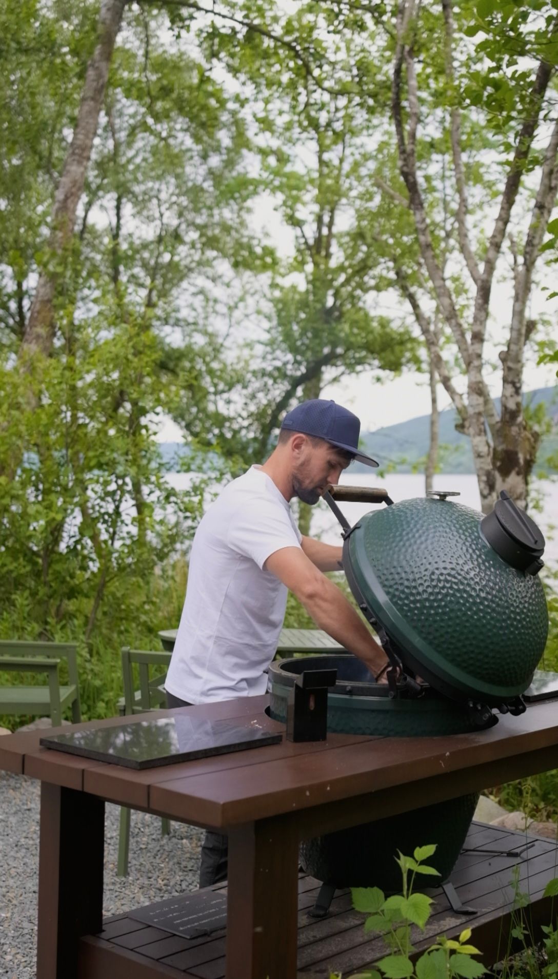 Man in white shirt and cap prepares a green ceramic grill on a wooden table outdoors.