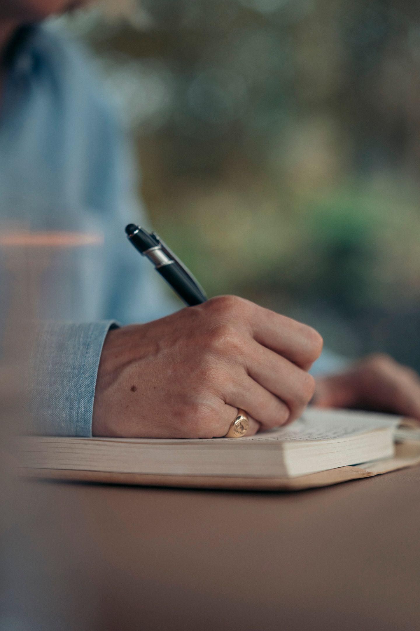 Person's hand writing in a notebook with a pen. Light blue shirt, gold ring. Beige table and book.