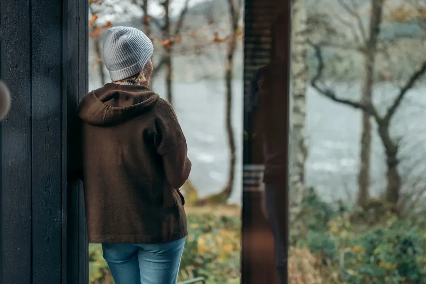 A guest looking out at a scenic loch view from a large window inside a cozy, modern woodland cabin.