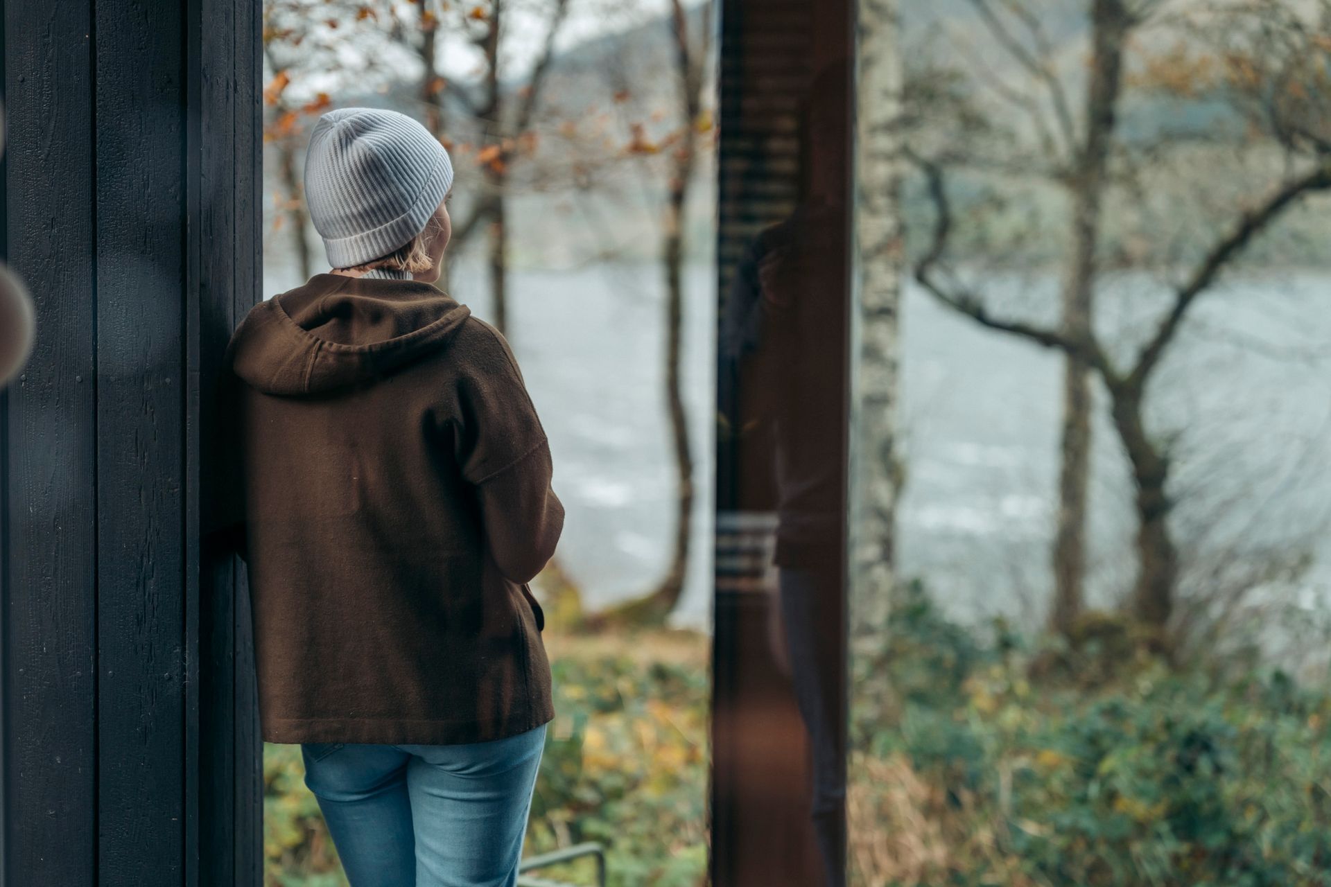 Person with gray hair, wearing a brown jacket, looks out at a lake and trees through a window.