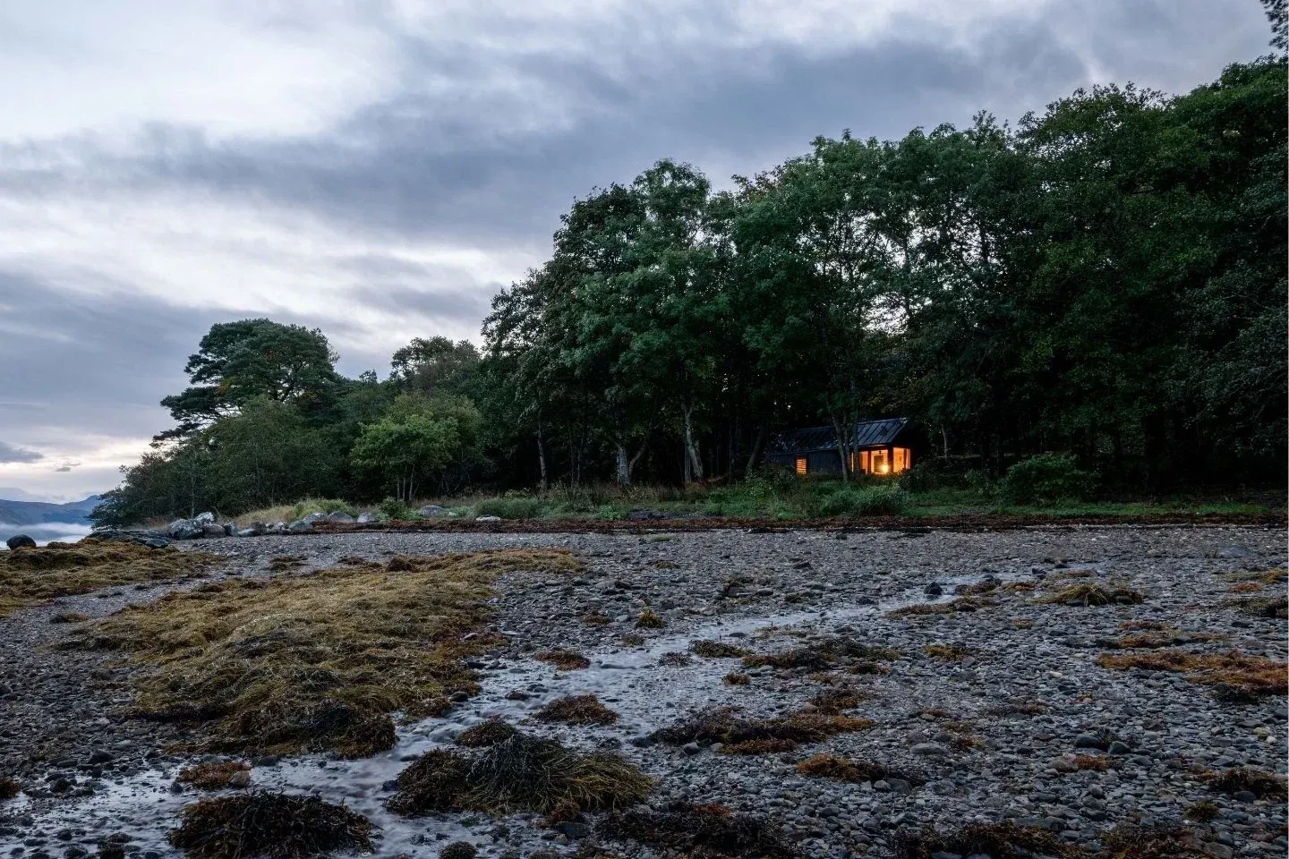 A small cabin with glowing lights at dusk situated on the rocky shores of a peaceful Scottish loch.