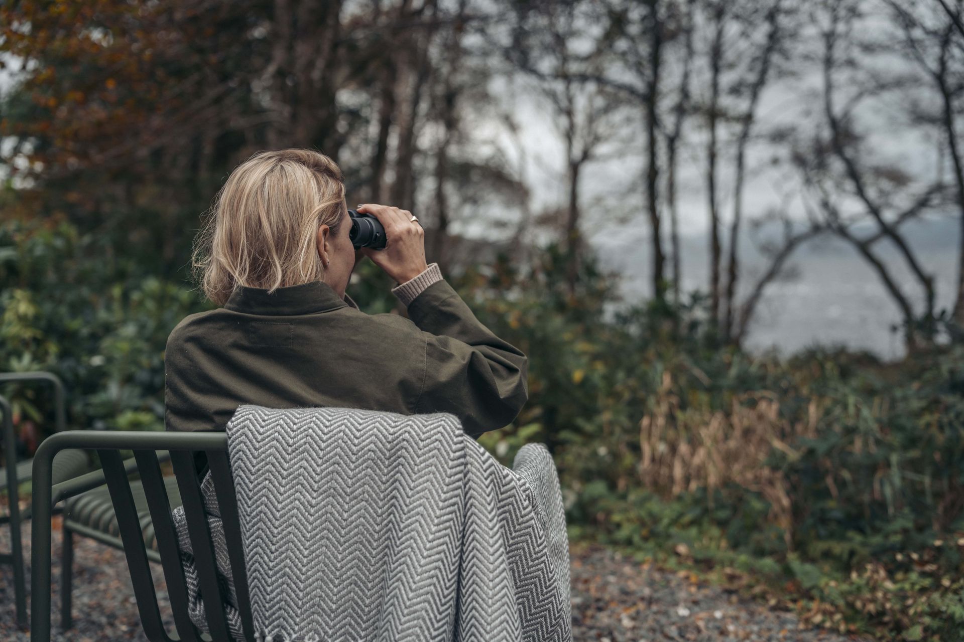 Person seated outdoors, using binoculars. Overcast day; trees and water visible in the background.