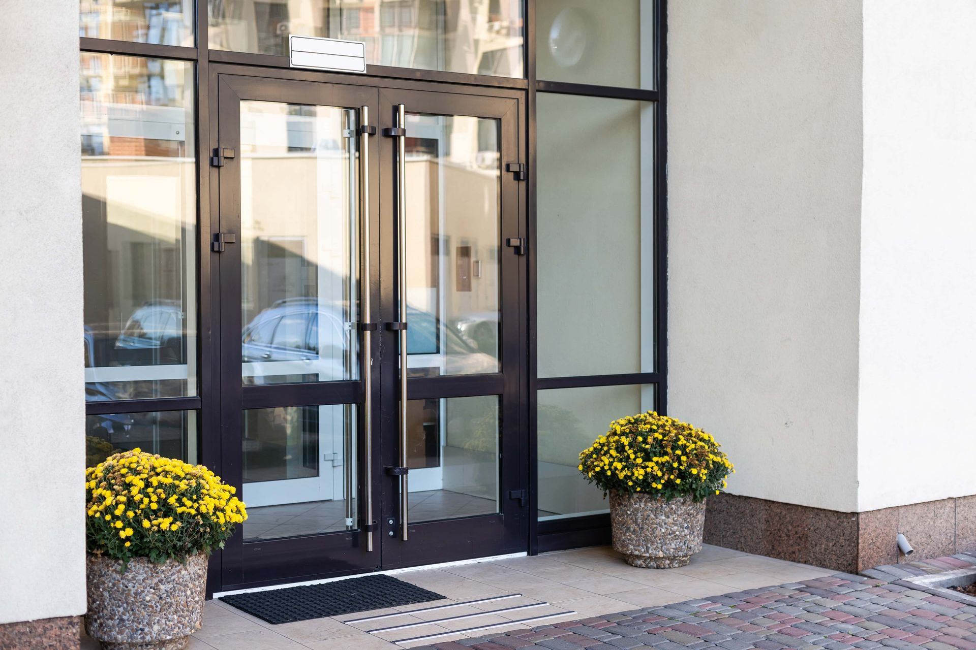 Double glass doors with dark frames, flanked by potted yellow flowers, in a building entrance.