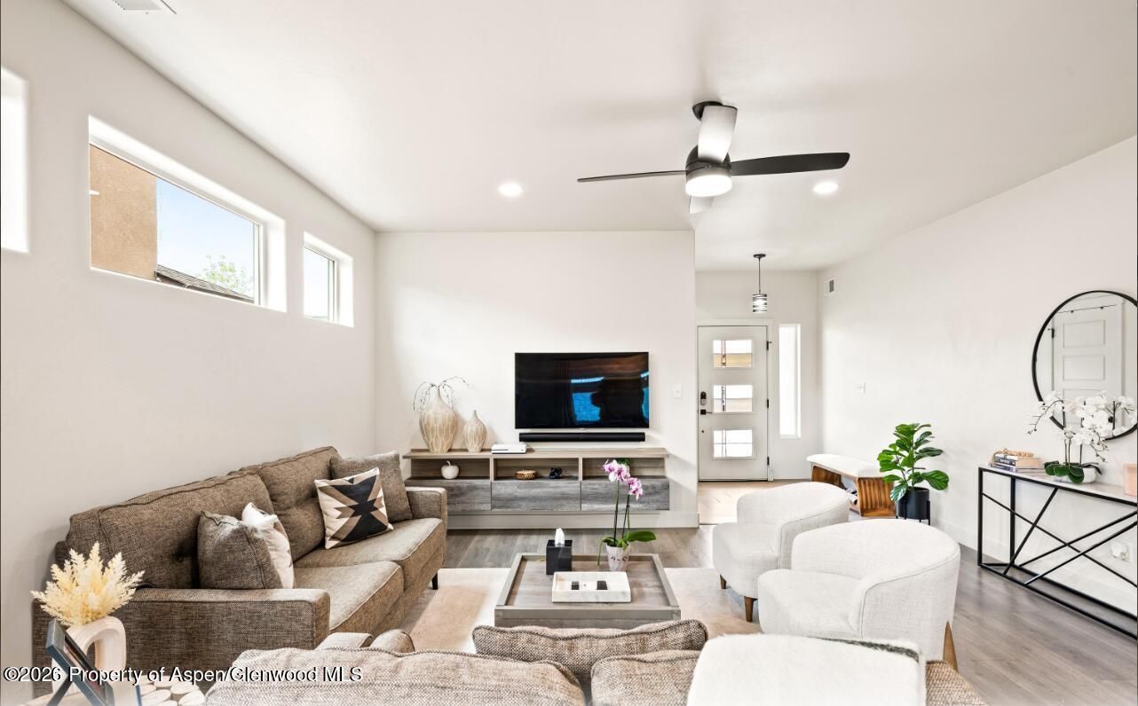 Bright modern living room with brown sectional, white chair, TV, coffee table, and ceiling fan.