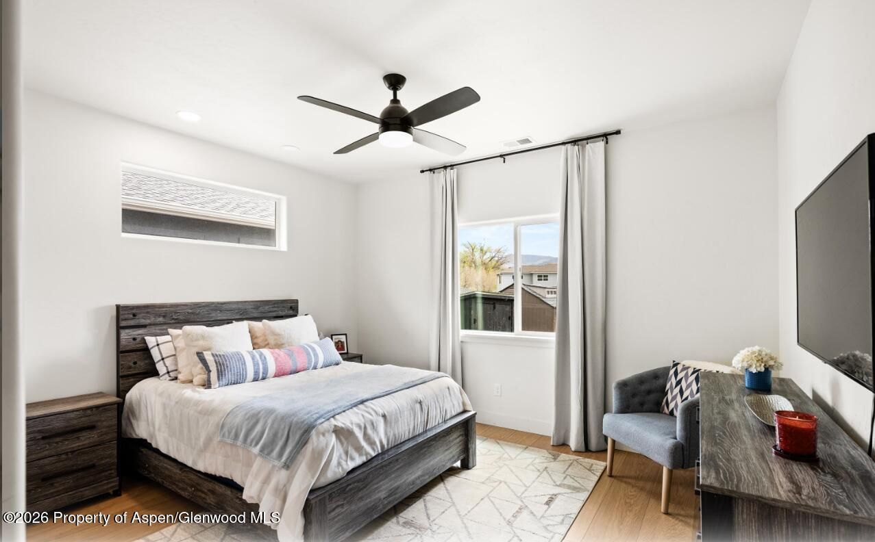 Bright bedroom with a bed, ceiling fan, window, chair, and TV on a white wall.