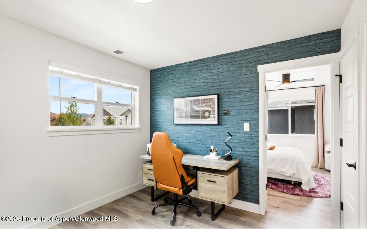 Small home office with teal accent wall, orange chair, and desk beside an open doorway to a bedroom.