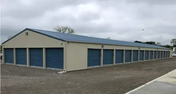 Exterior view of storage units, tan building with blue doors and roof, gravel parking.