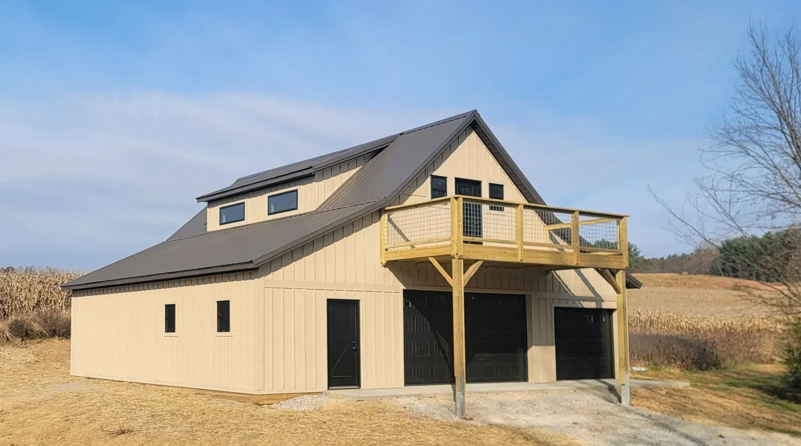 Tan barn-style building with dark gray roof, deck, and black garage doors, set in a field.