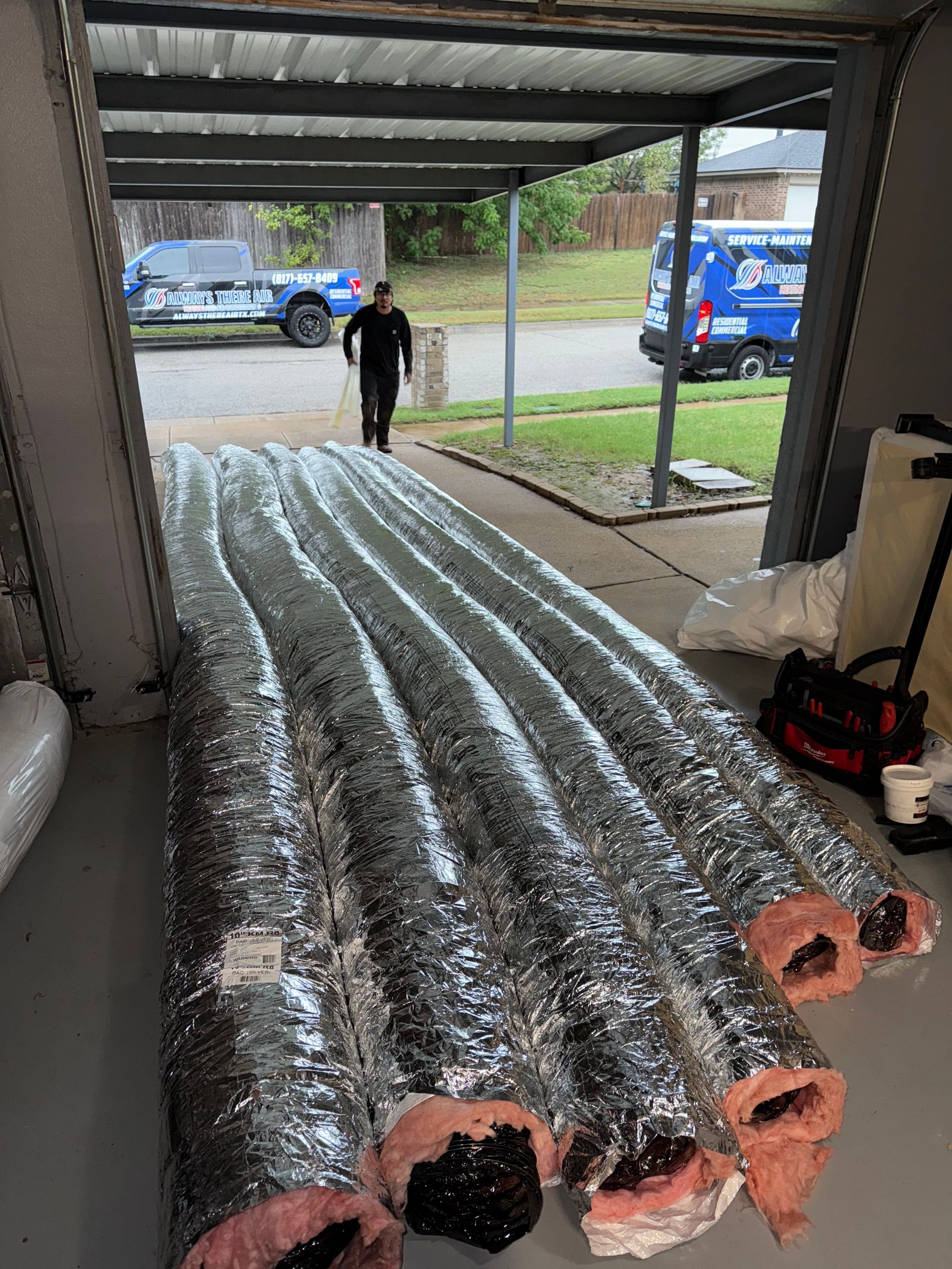 A person walking into a garage with silver air ducts, a trailer and service van visible outside.