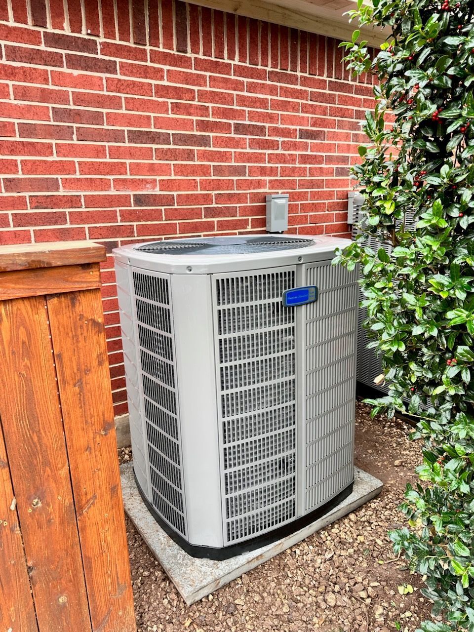 Air conditioner unit against a brick wall, next to a wooden fence and greenery.