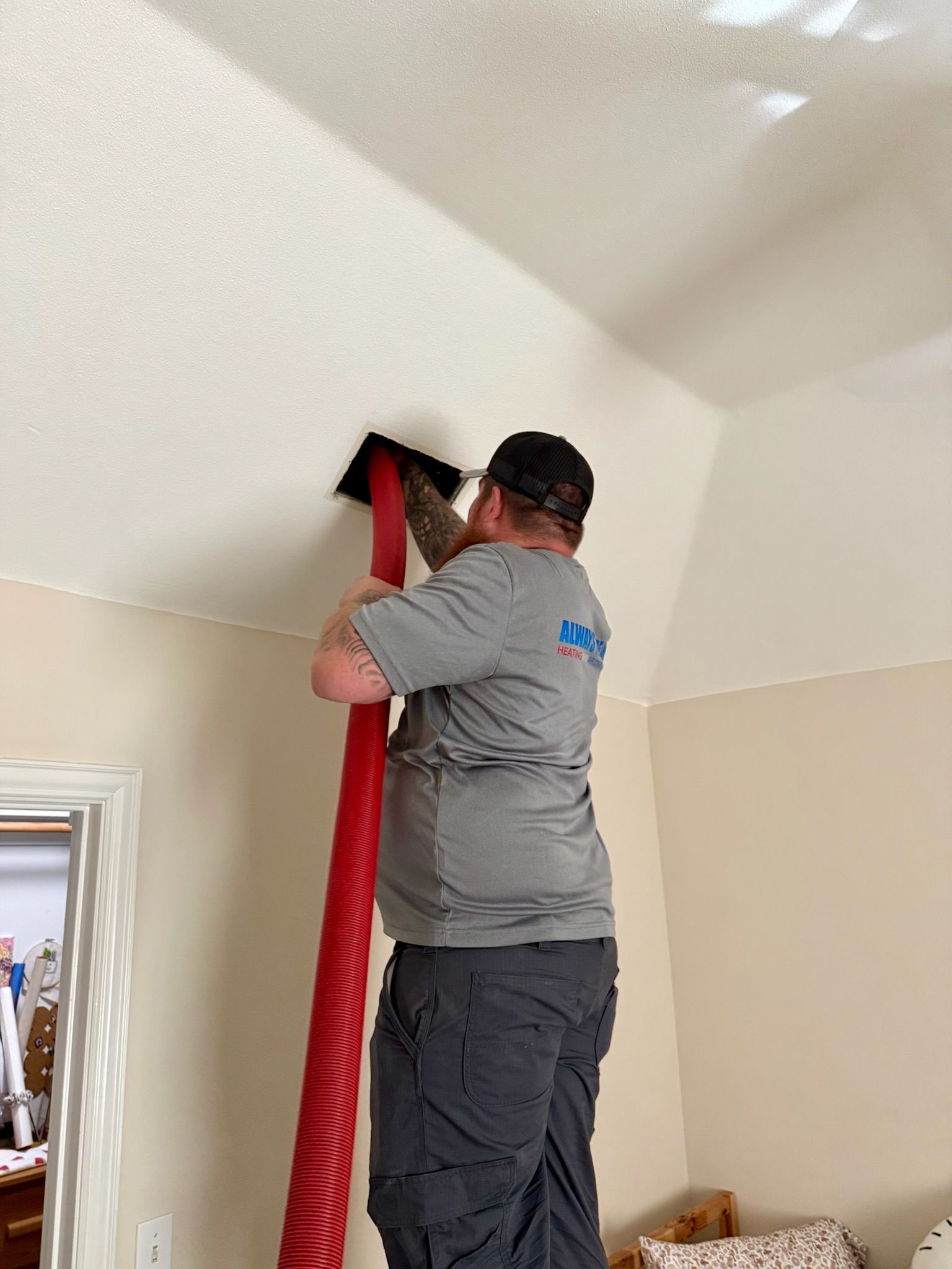 Man on a ladder cleaning ceiling vent with a red hose. Interior shot, beige walls.