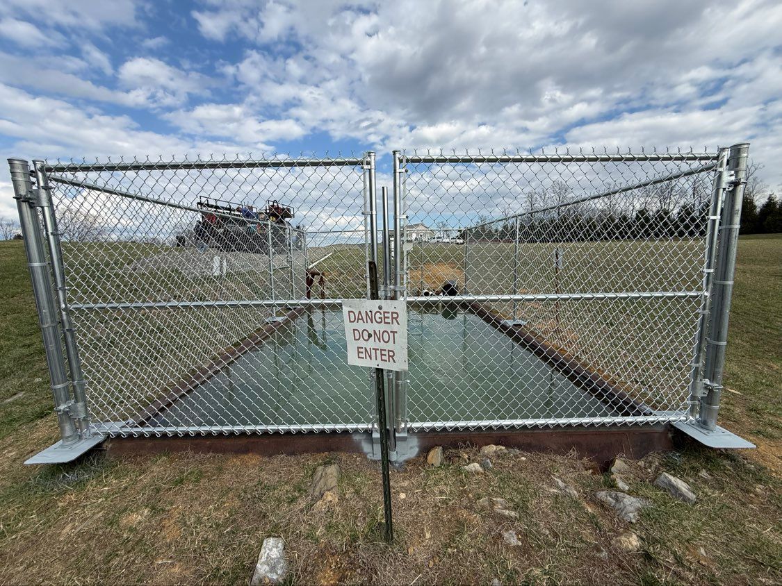 Chain-link gate blocking a narrow paved path, with a warning sign in front and fields on both sides.