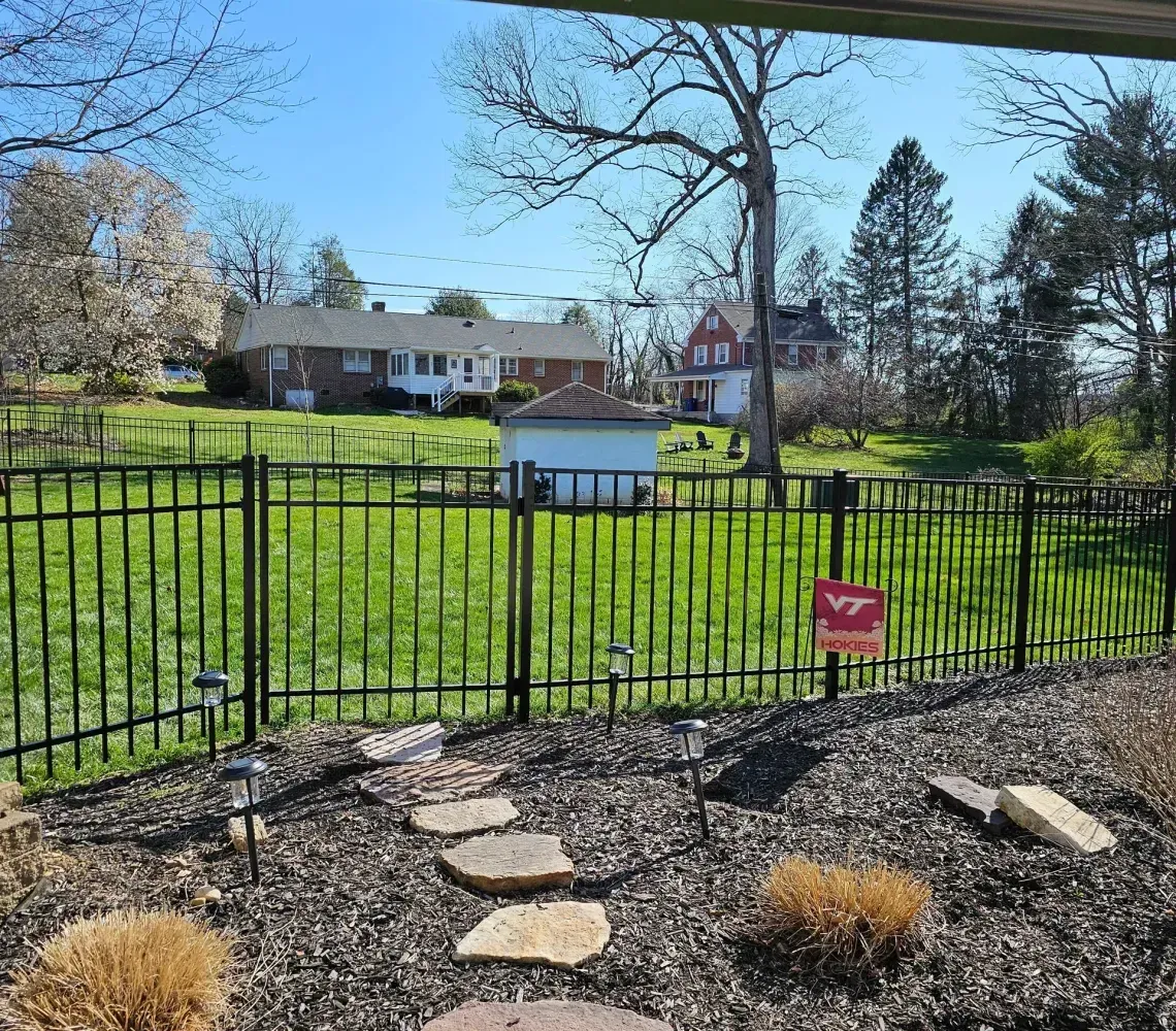 Black metal fence around a backyard garden, with a house and blue sky in the background.