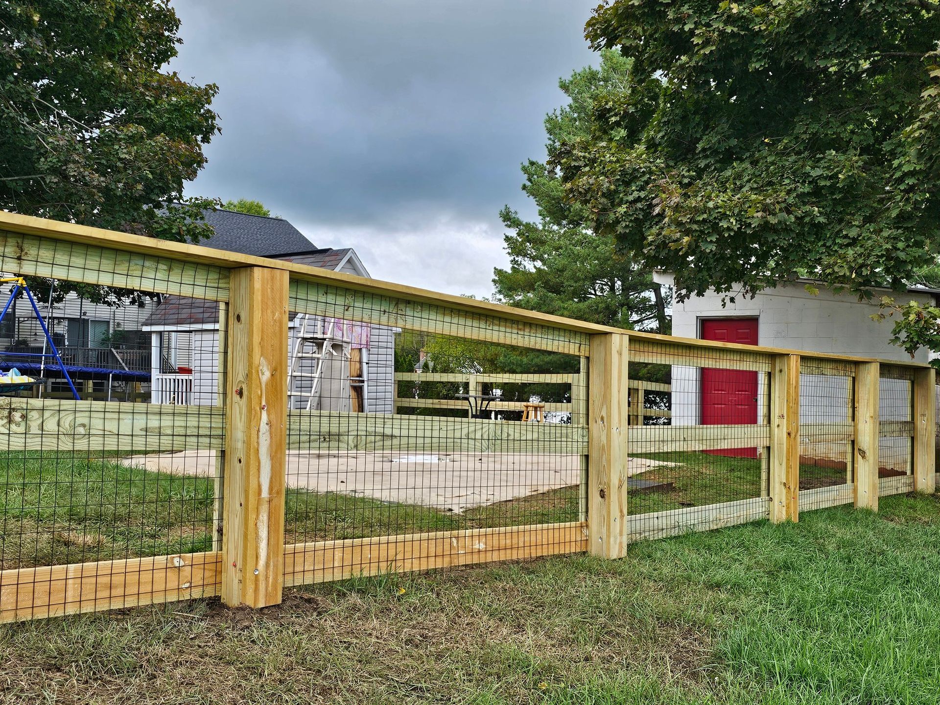 Wooden fence along a grassy yard with buildings and trees under a cloudy sky