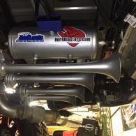 Under-car view of a silver aftermarket exhaust system with blue and red branded canisters in a garage