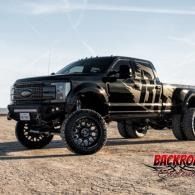 Lifted black pickup truck with oversized off-road tires parked on a dirt lot under a blue sky