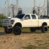 White lifted pickup truck parked on dirt near sailboat masts, with large black off-road tires.