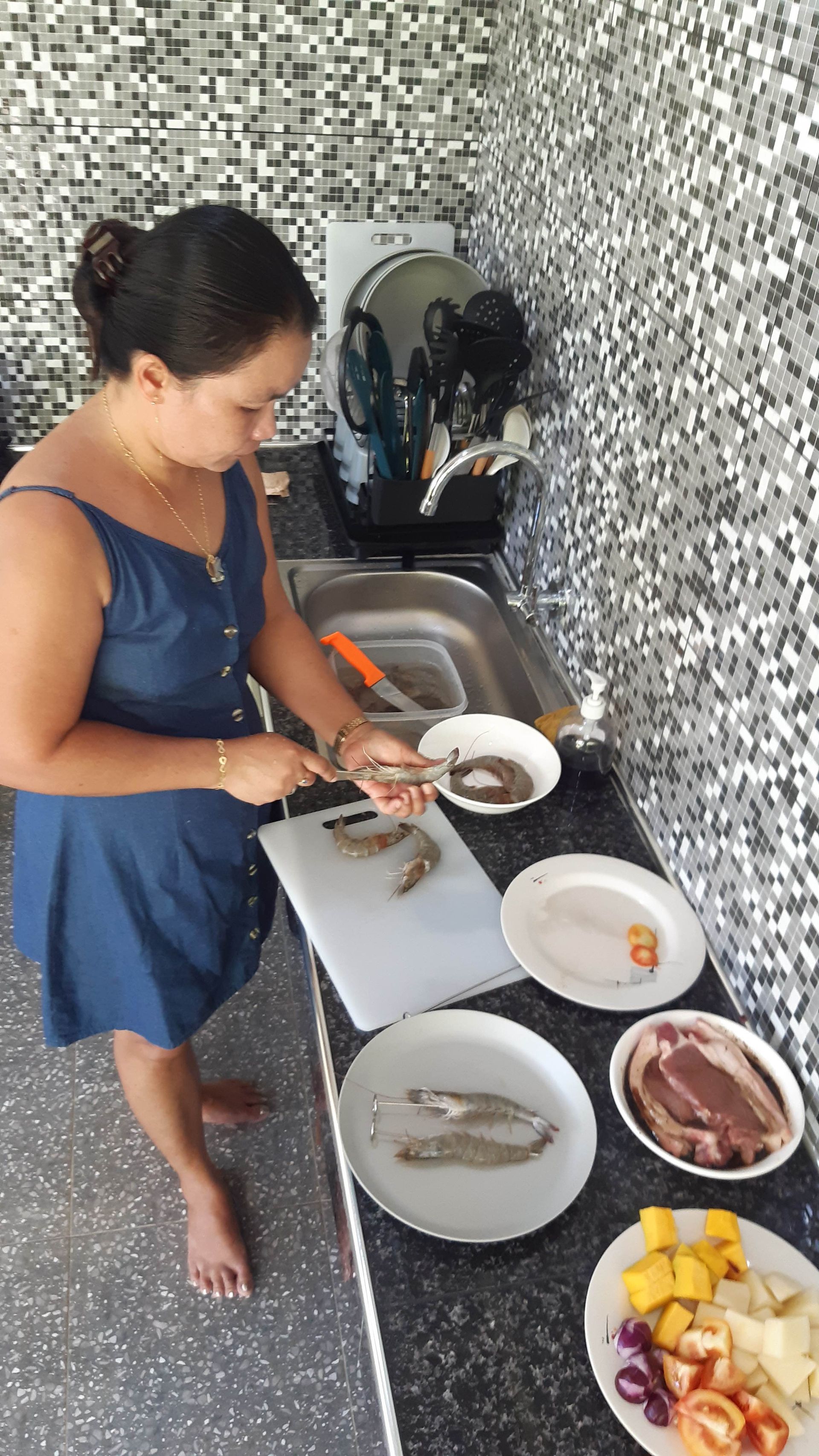 Woman in blue dress prepping shrimp in a kitchen; ingredients & sink visible.