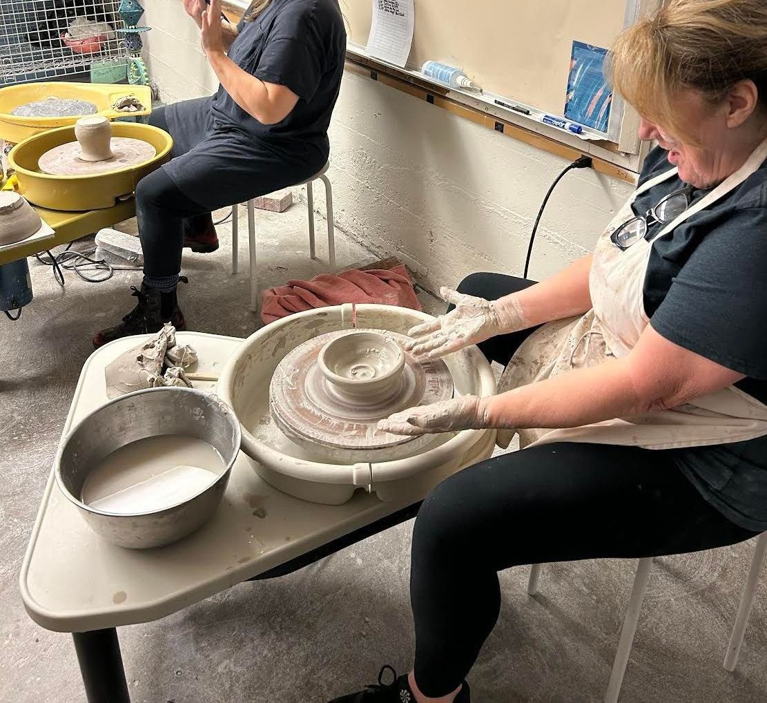 A woman is sitting at a table making a bowl on a pottery wheel.