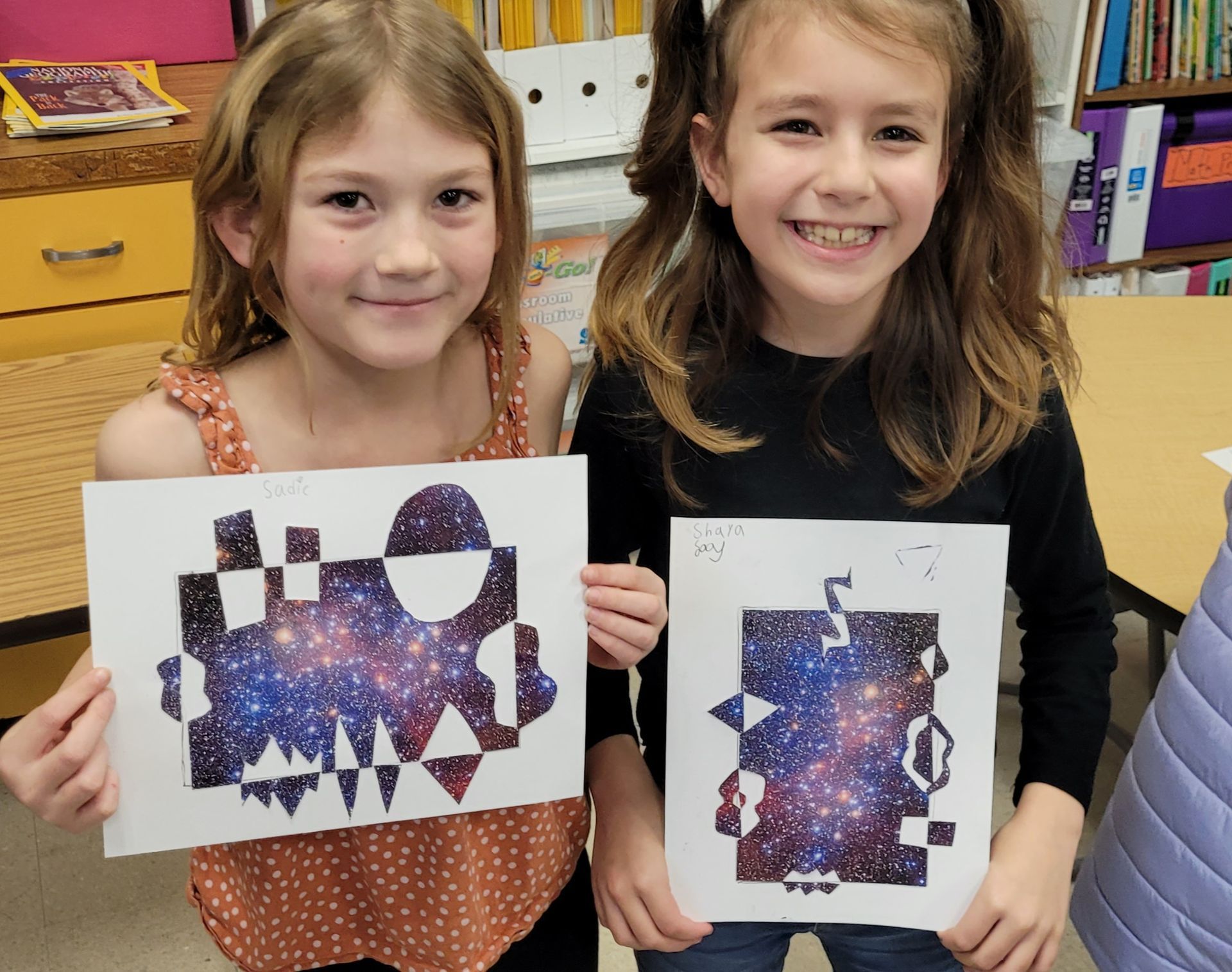 Two young girls are holding up their artwork in a classroom.