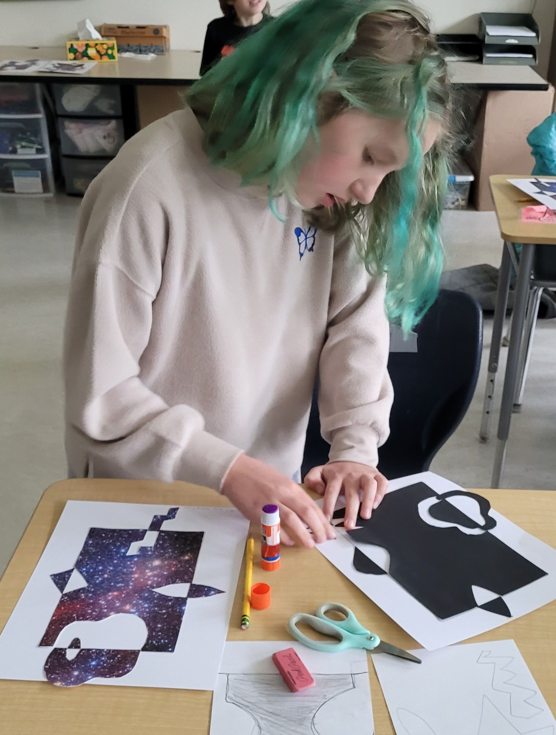A girl with green hair is sitting at a table with scissors and glue