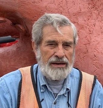 A man with a beard and an orange vest is standing in front of a red wall.