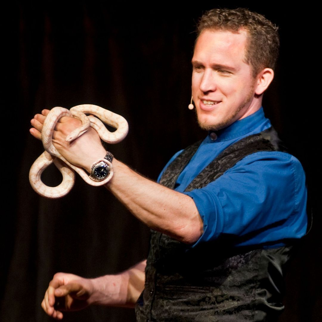 A man in a blue shirt is holding a snake in his hand FOI - Magic - Spring Break