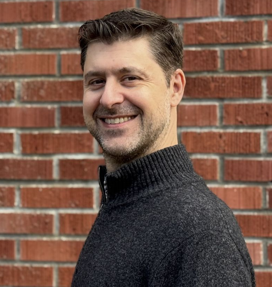 A woman with long blonde hair is smiling and wearing a black shirt and earrings.