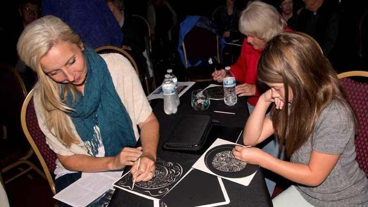 A woman and a girl are sitting at a table drawing on a piece of paper.