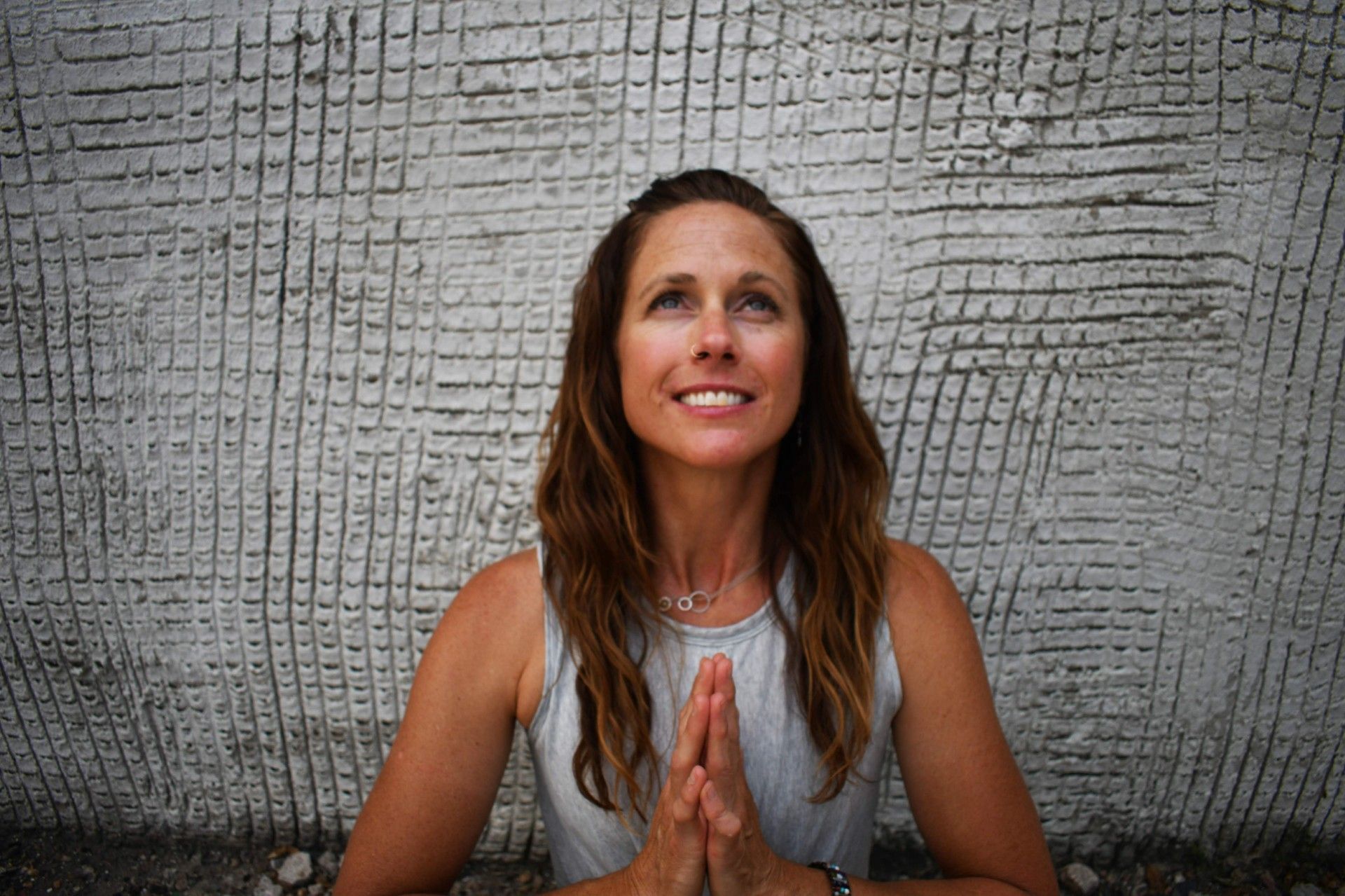 A woman is sitting in front of a brick wall with her hands folded and smiling.