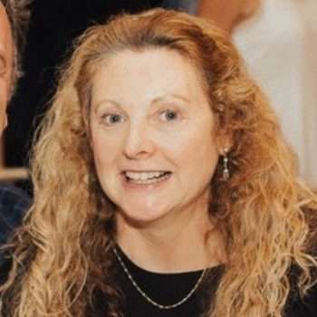 A woman with long blonde hair is smiling and wearing a black shirt and earrings.