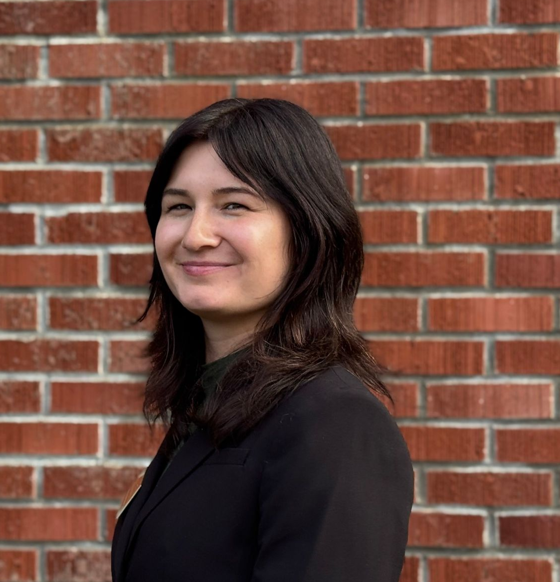 A woman wearing a black shirt with a flower pattern and a black blazer on is smiling.