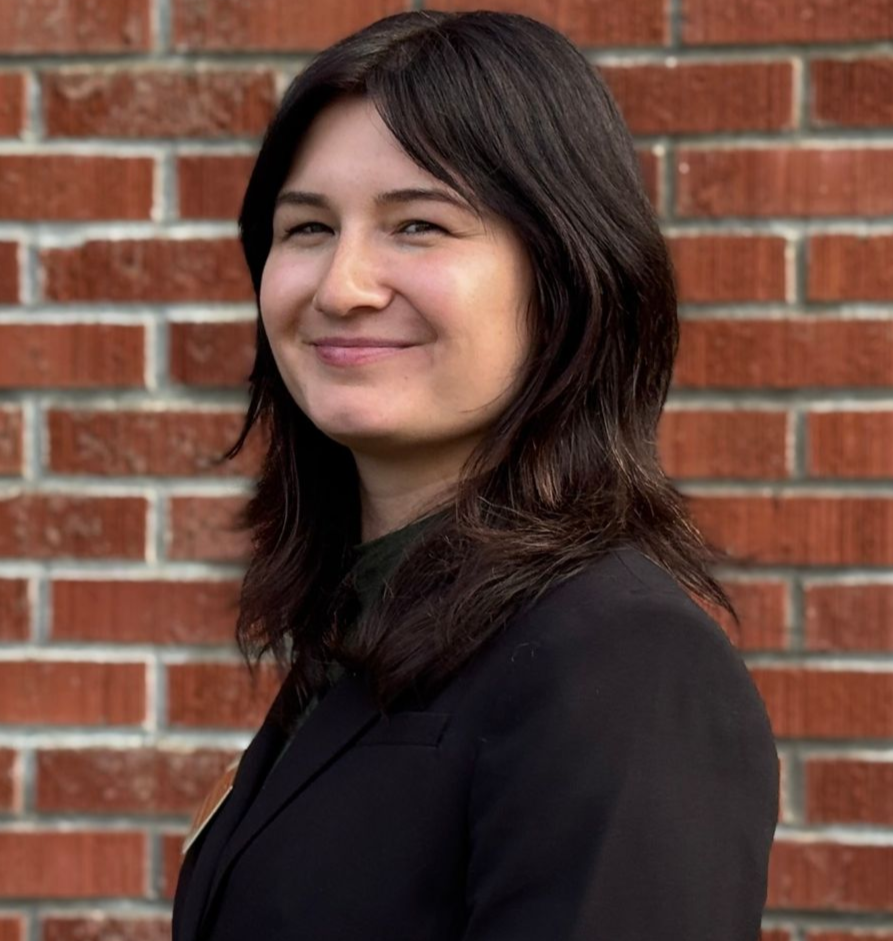 A woman wearing a black shirt with a flower pattern and a black blazer on is smiling.