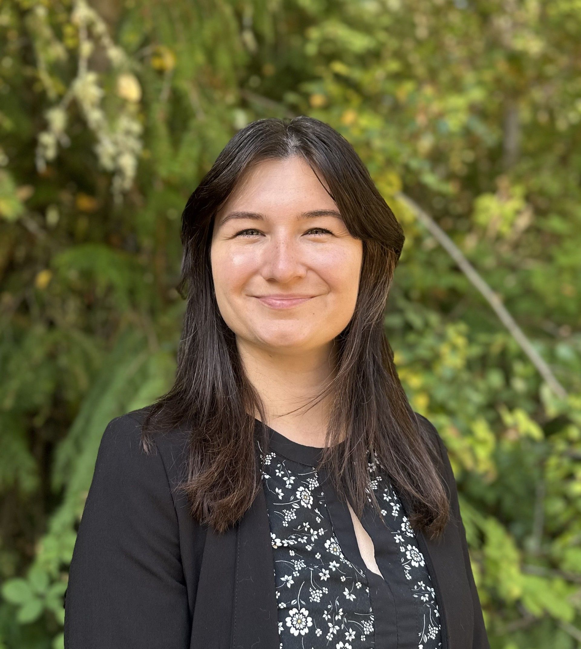 A woman wearing a black shirt with a flower pattern and a black blazer on is smiling.