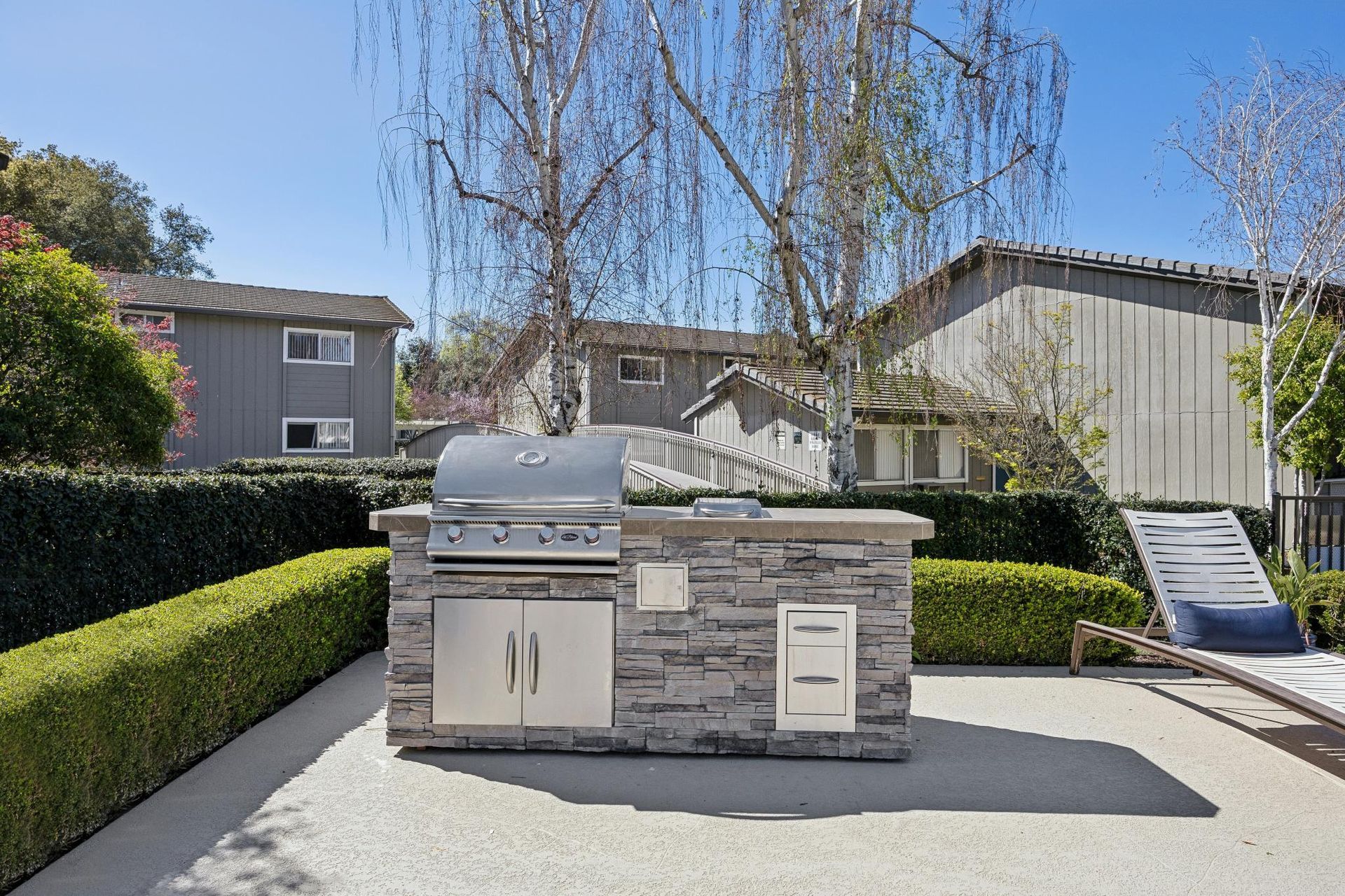 Outdoor kitchen with stone island, stainless steel grill, and cabinetry in a backyard setting.