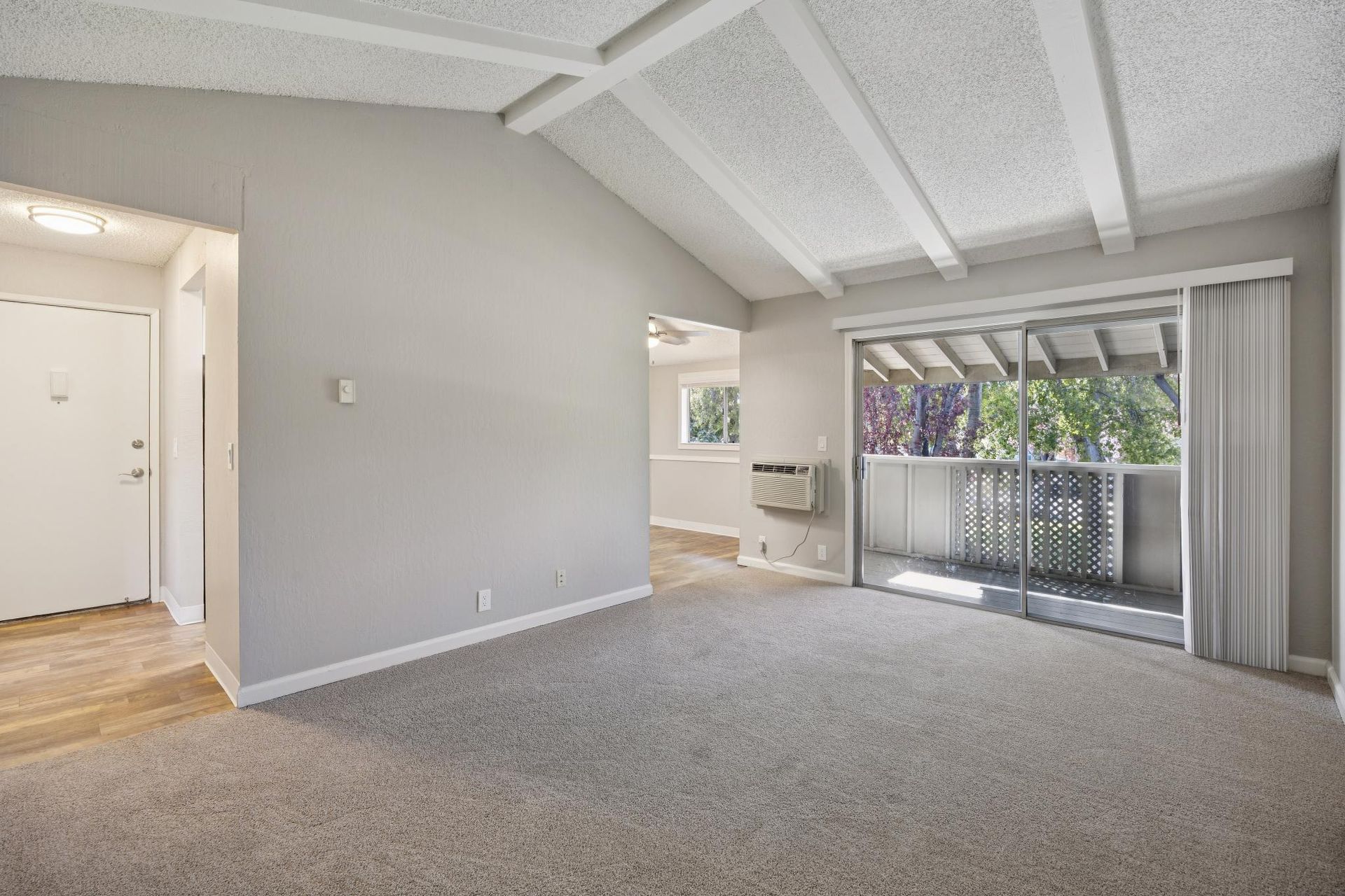 Empty living room with gray carpet, vaulted ceiling, and sliding glass door to balcony.