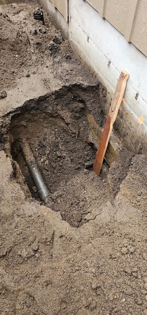 Hole dug in soil near a building, revealing pipes and a wooden stake.