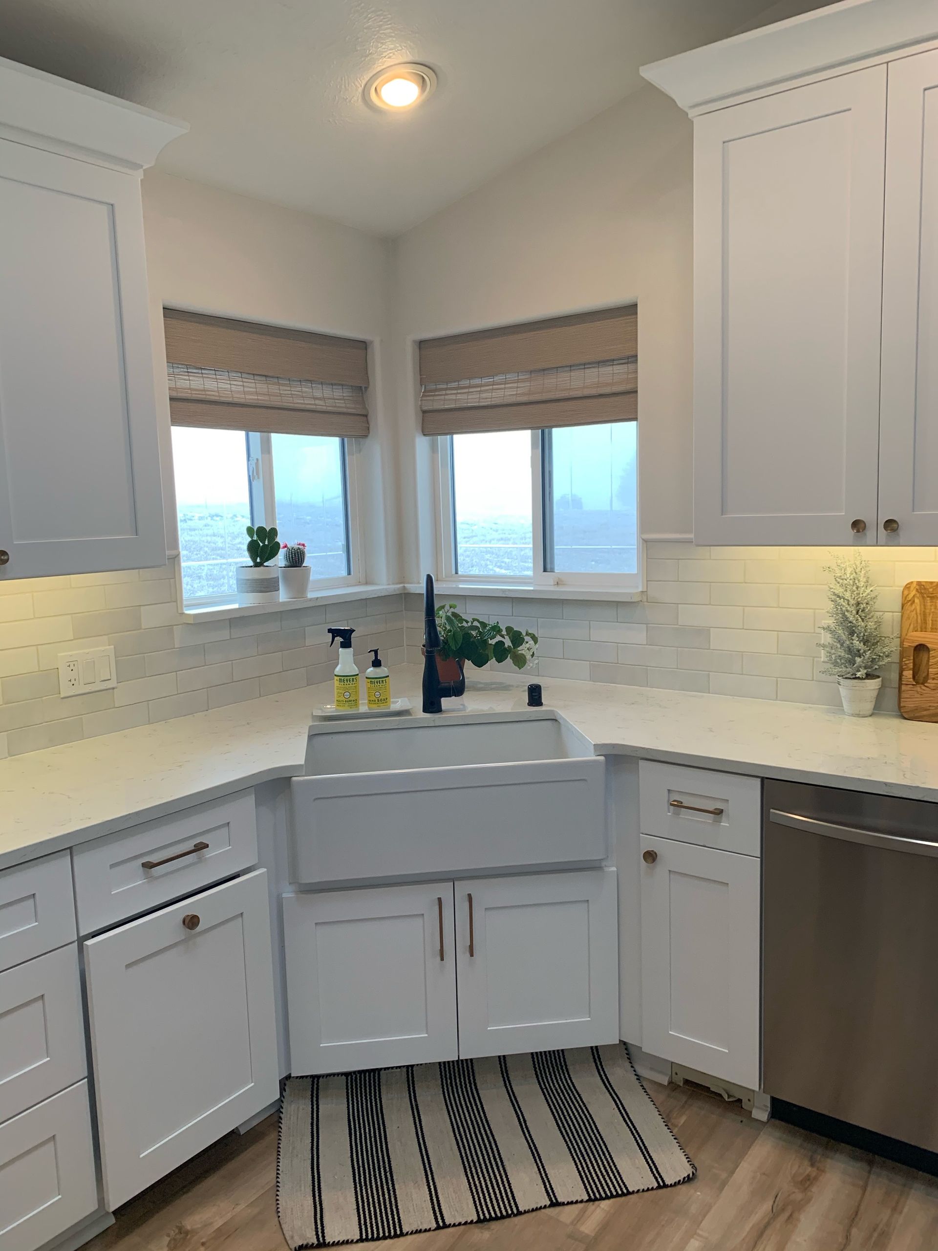 White kitchen corner with farmhouse sink, windows, and cabinets. Black and white rug, and light countertops.