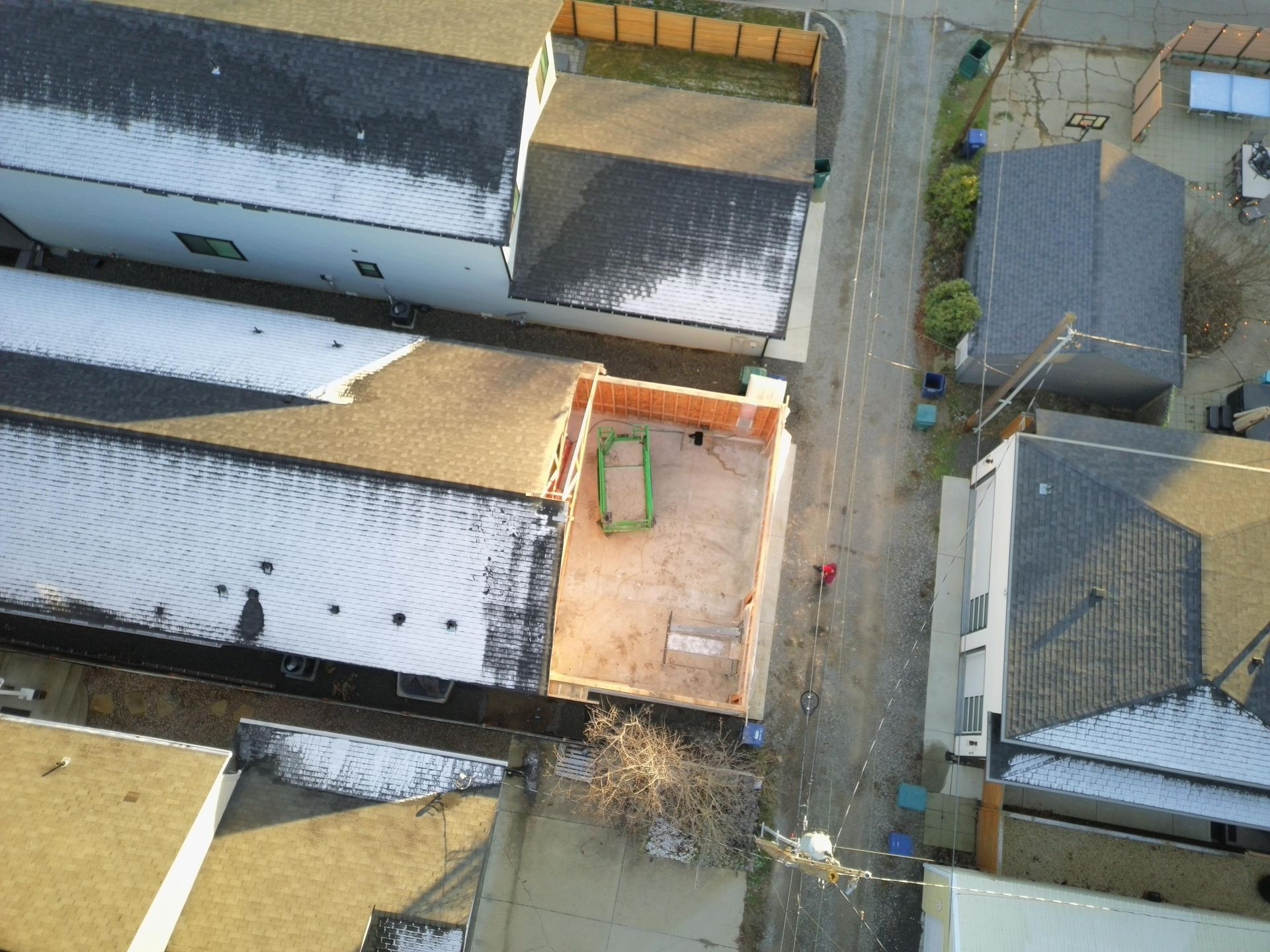 Aerial view: construction site with exposed wooden framing, surrounded by houses and a street. A person walks on the road.