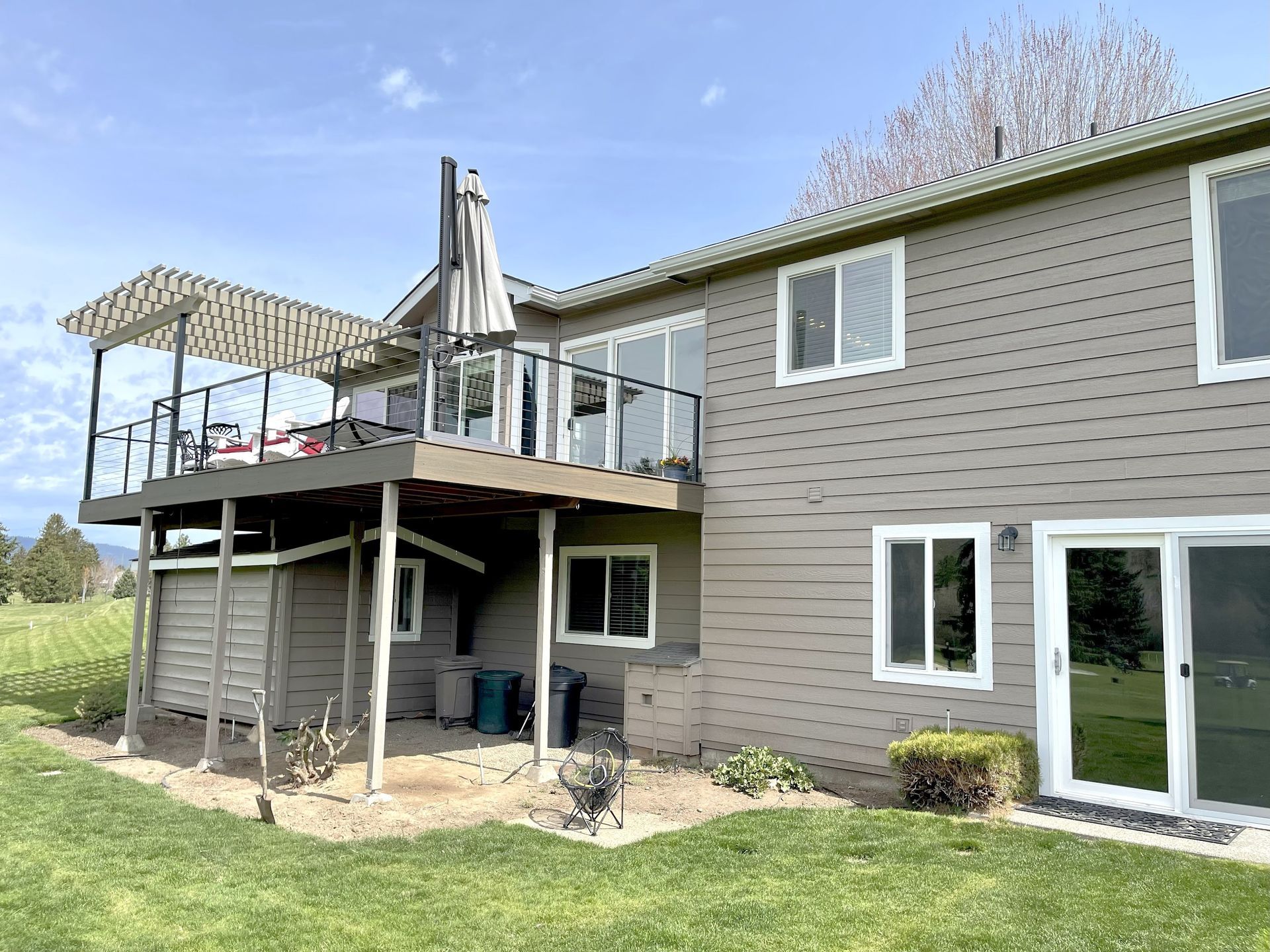 Two-story house with deck. Beige siding, white-framed windows, glass railings. Green lawn and blue sky background.