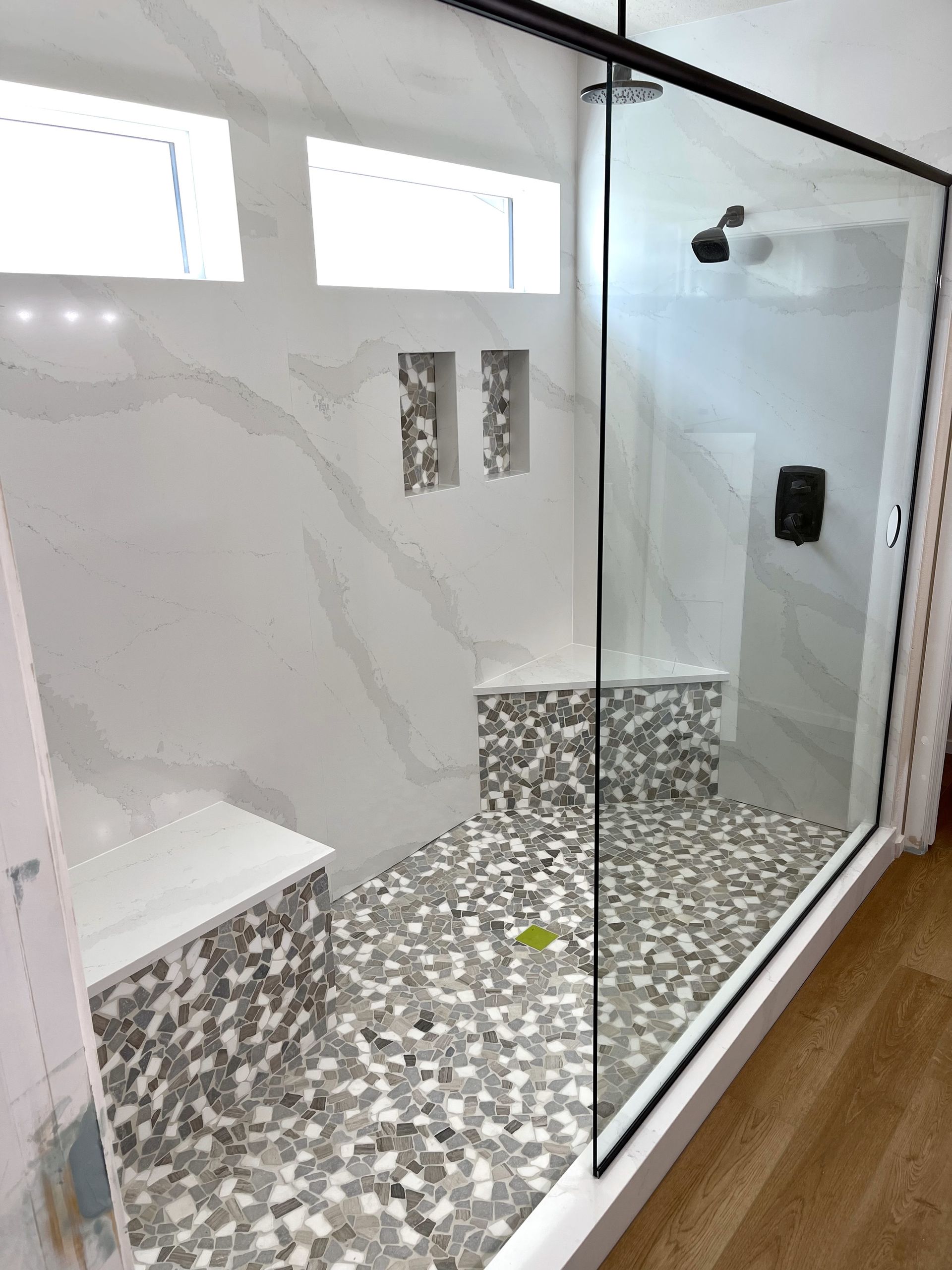 Spacious, modern shower with glass door, marble-like tiles, built-in bench, and pebble floor.
